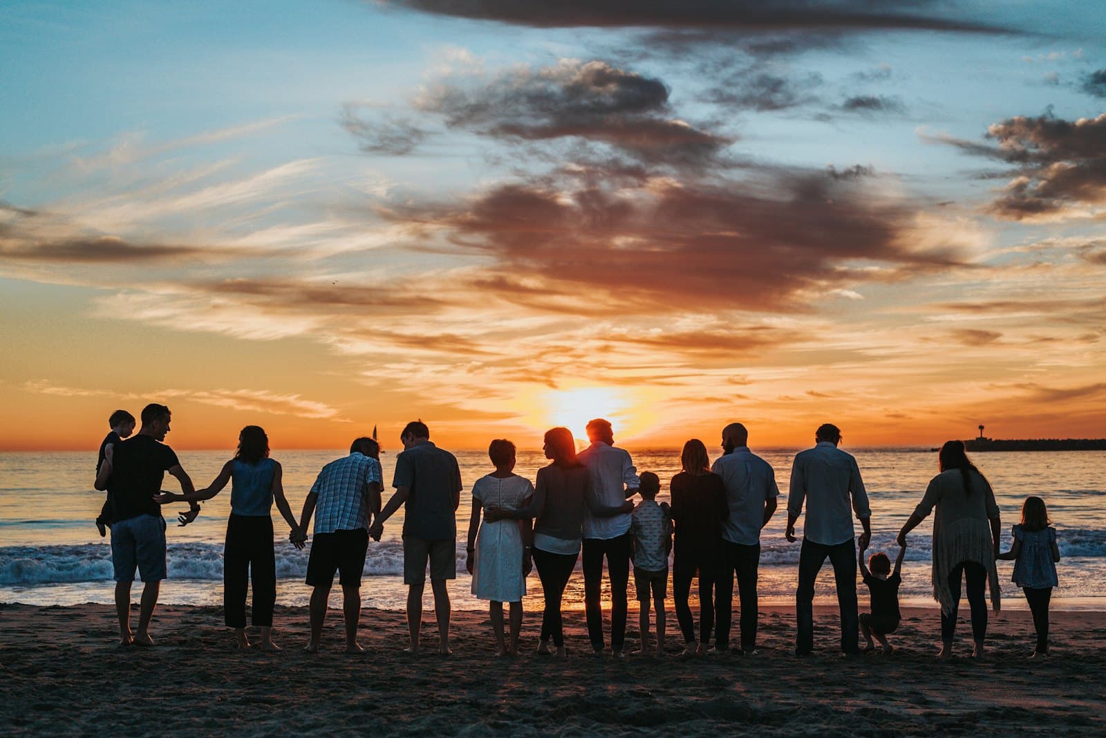 People of all ages together at the shore at sunset, holding on to each other.